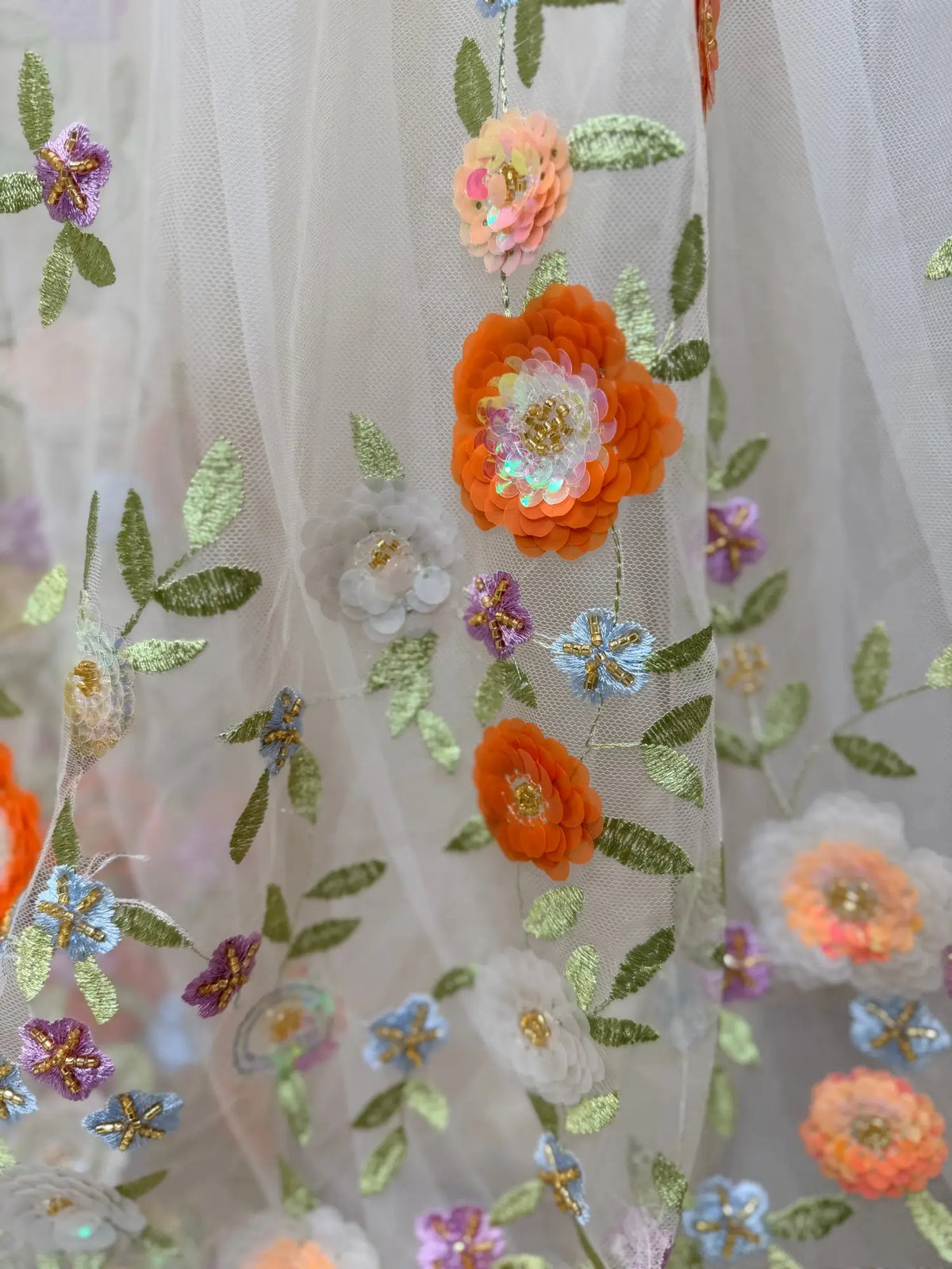 Sheer tulle wedding dress fabric with vibrant multicolor floral embroidery, sequins, and delicate green leaf detailing.