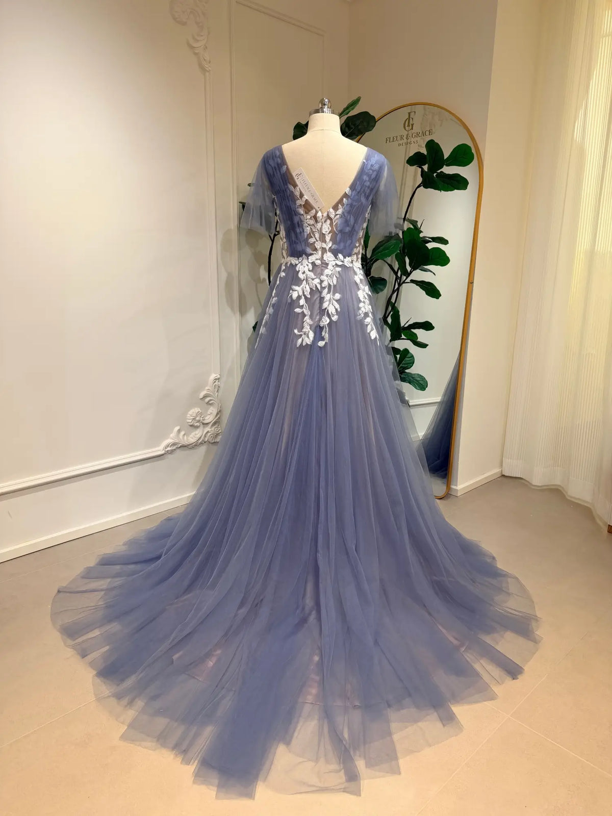Dusty blue A-line wedding dress with white floral lace appliqués, flutter sleeves, and a flowing tulle skirt on a mannequin.