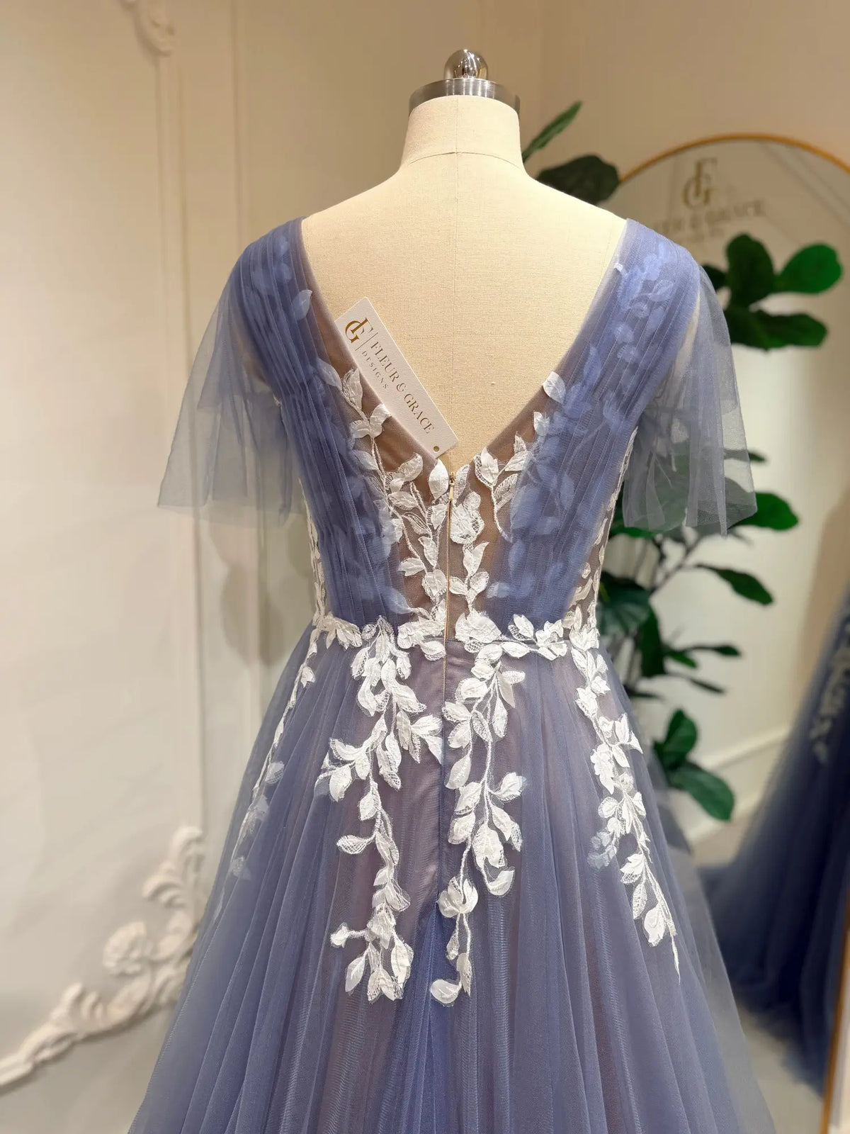 Dusty blue A-line wedding dress with off-white floral lace, illusion V-back, and romantic flutter sleeves on a mannequin.
