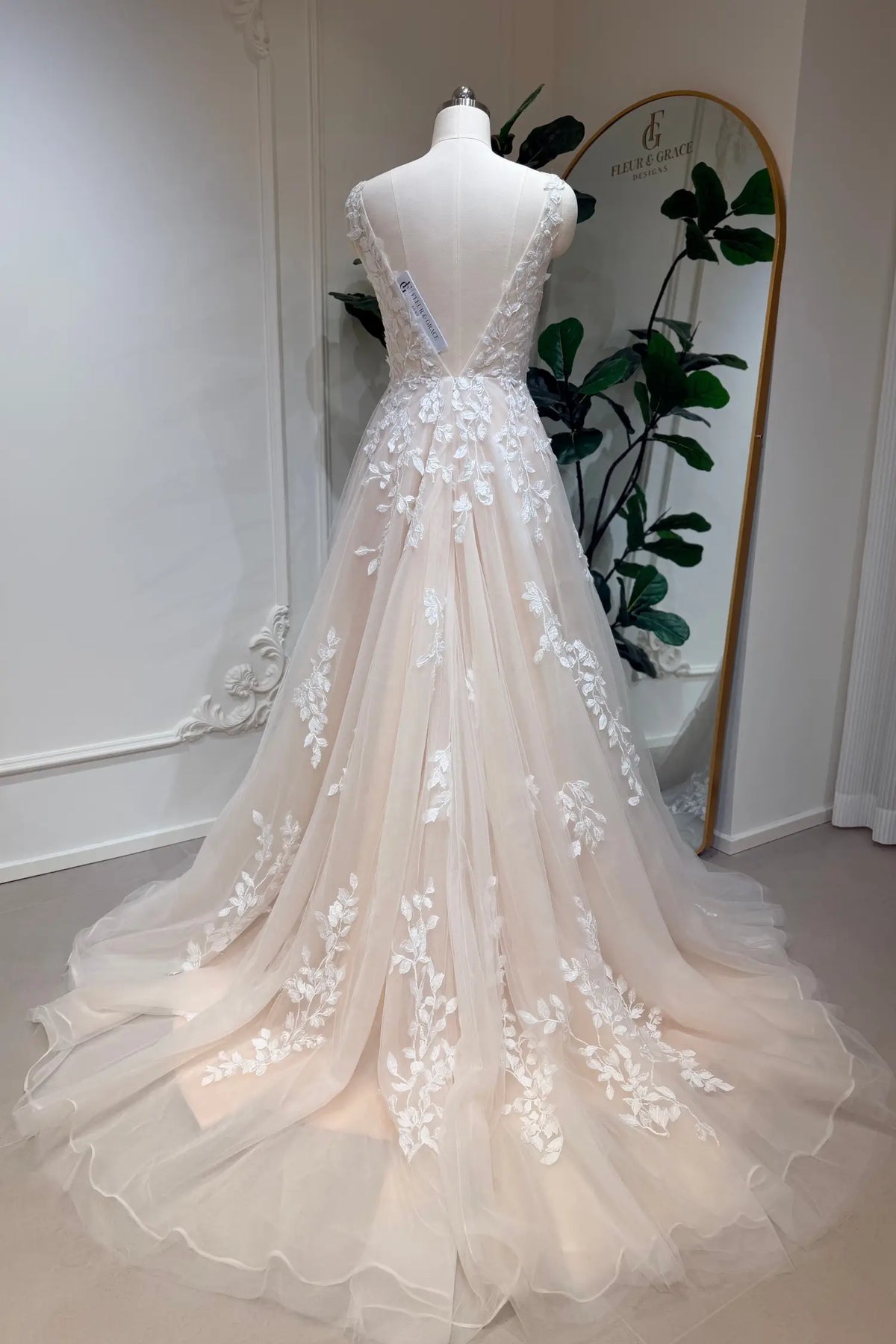 Champagne A-line wedding dress with floral lace appliqué, deep V-back, and flowing tulle skirt on a mannequin.