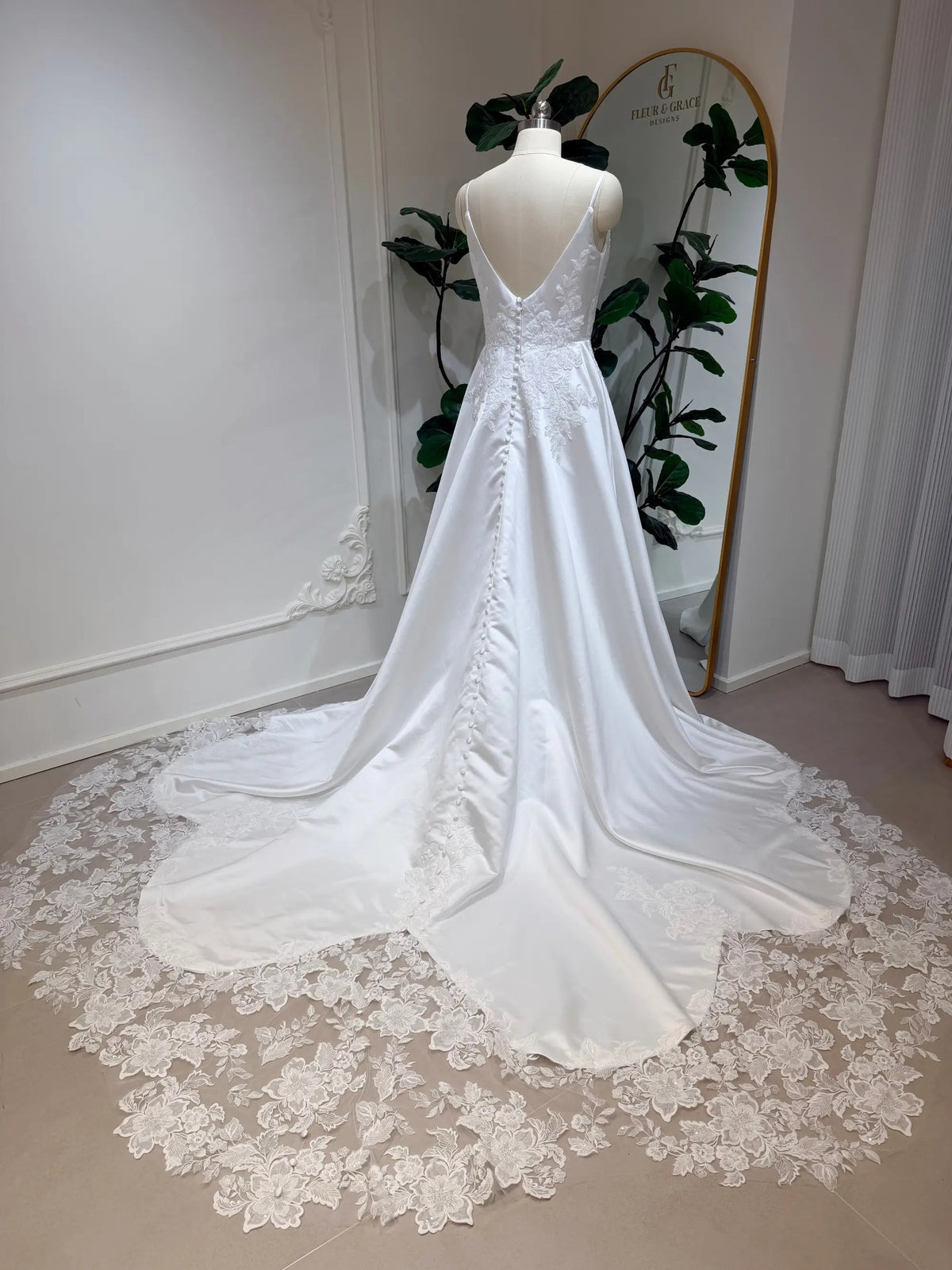 White satin A-line wedding dress with a deep V-back, button details, and a floral lace appliquéd train on a mannequin.
