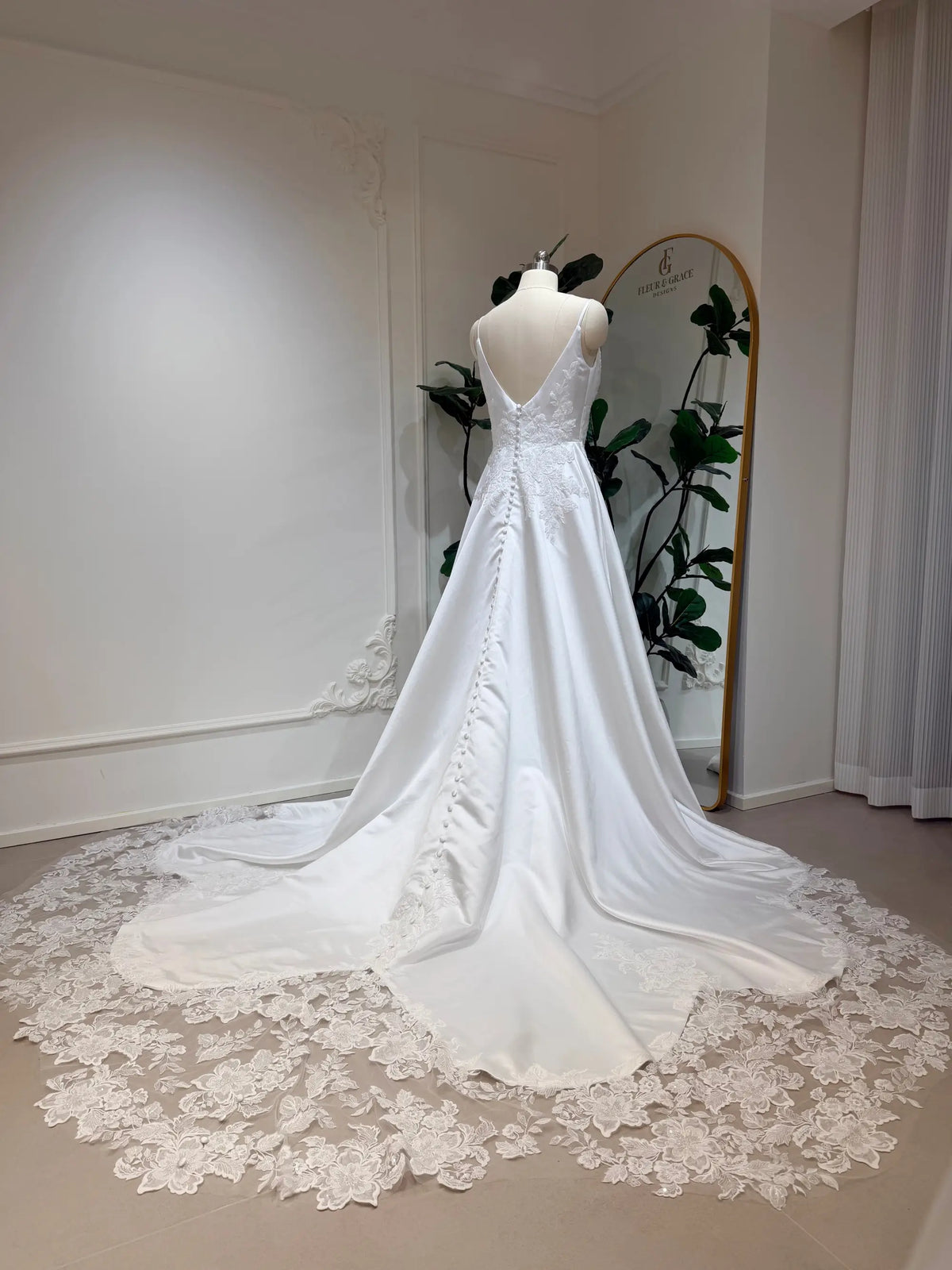 Satin A-line wedding dress with a deep V-back, button details, and a floral lace appliquéd train on a mannequin.