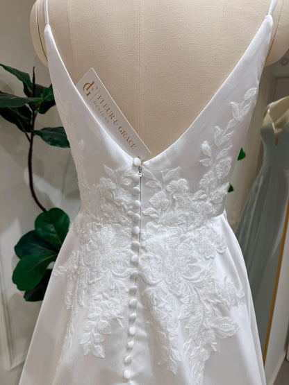 White satin A-line wedding dress with a deep V-back, delicate floral lace appliqué, and a row of elegant fabric buttons.