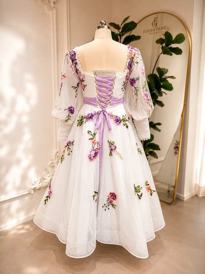 Tea-length wedding dress with lavender floral embroidery, puff sleeves, and a lace-up corset back on a white tulle skirt.
