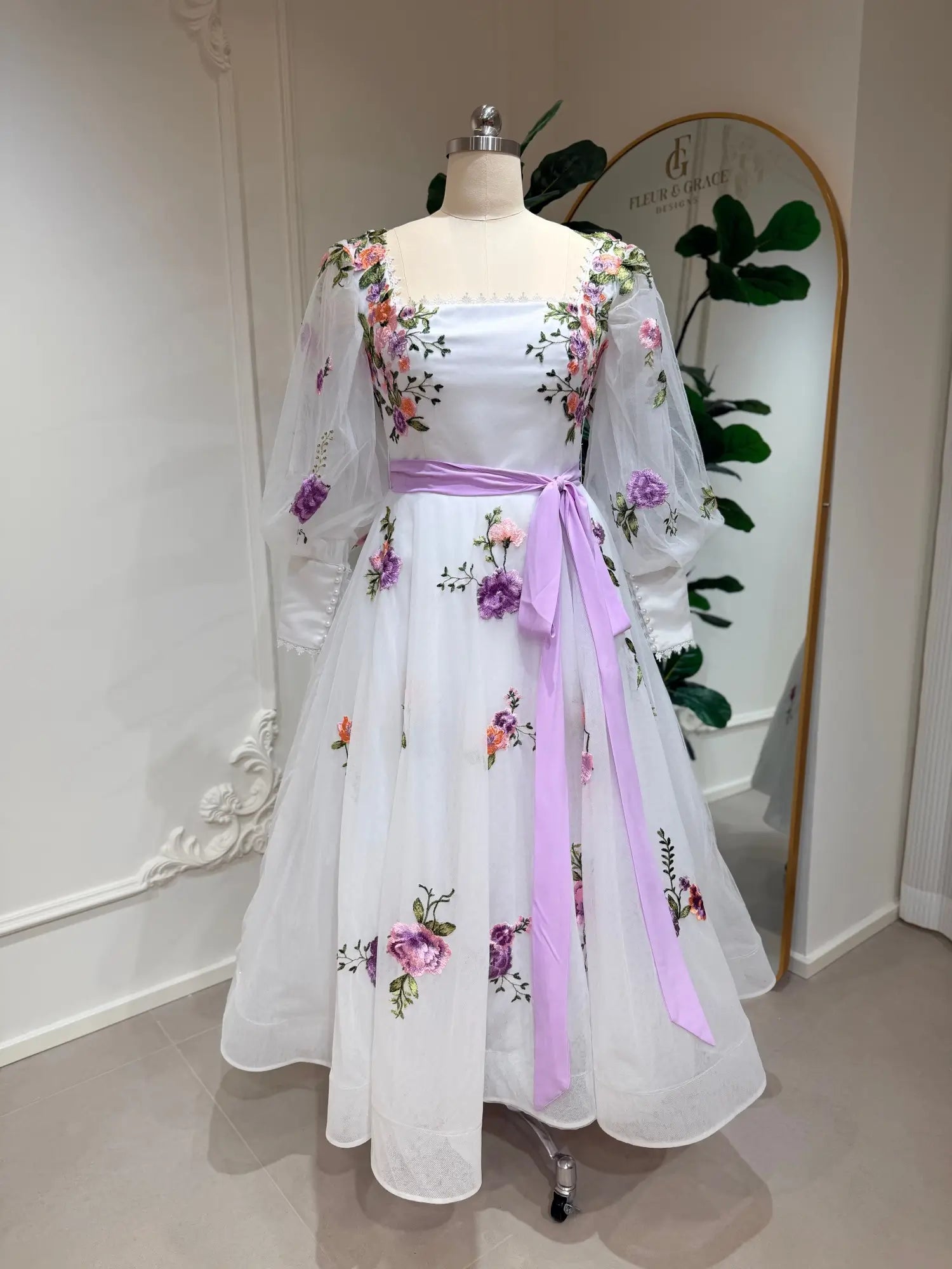 Tea-length wedding dress with lavender floral embroidery, puff sleeves, and a satin waist sash on a mannequin.