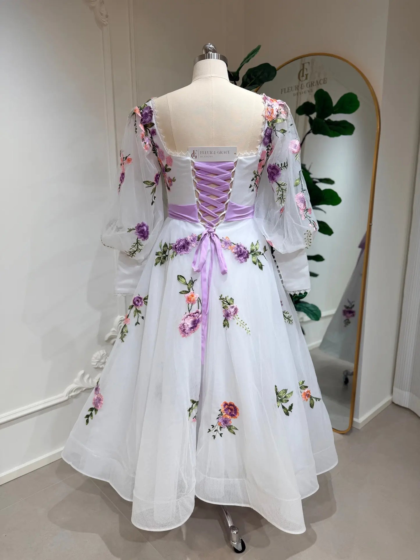 Tea-length wedding dress with lavender floral embroidery, puff sleeves, and a lavender lace-up corset back on a mannequin.