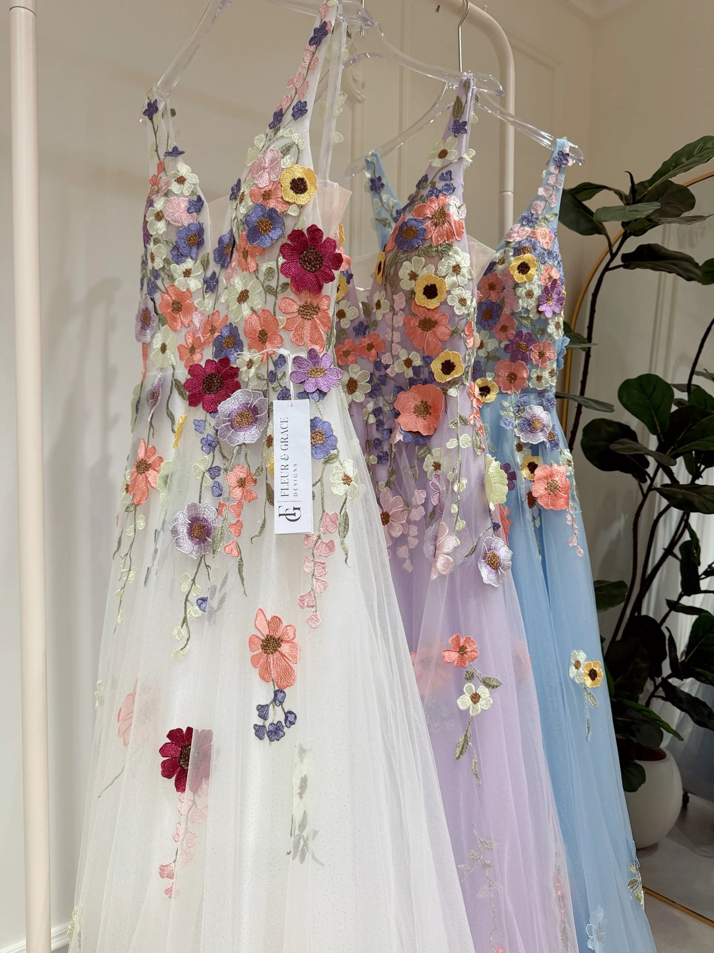 White, lavender, and blue A-line gowns featuring colorful wildflower embroidery, V-necklines, and sparkly mesh overlays.