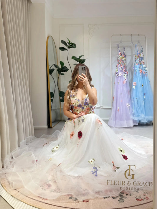 White A-line gown with colorful wildflower embroidery, sparkly mesh overlay, V-neckline, and a flowing floor-length skirt.