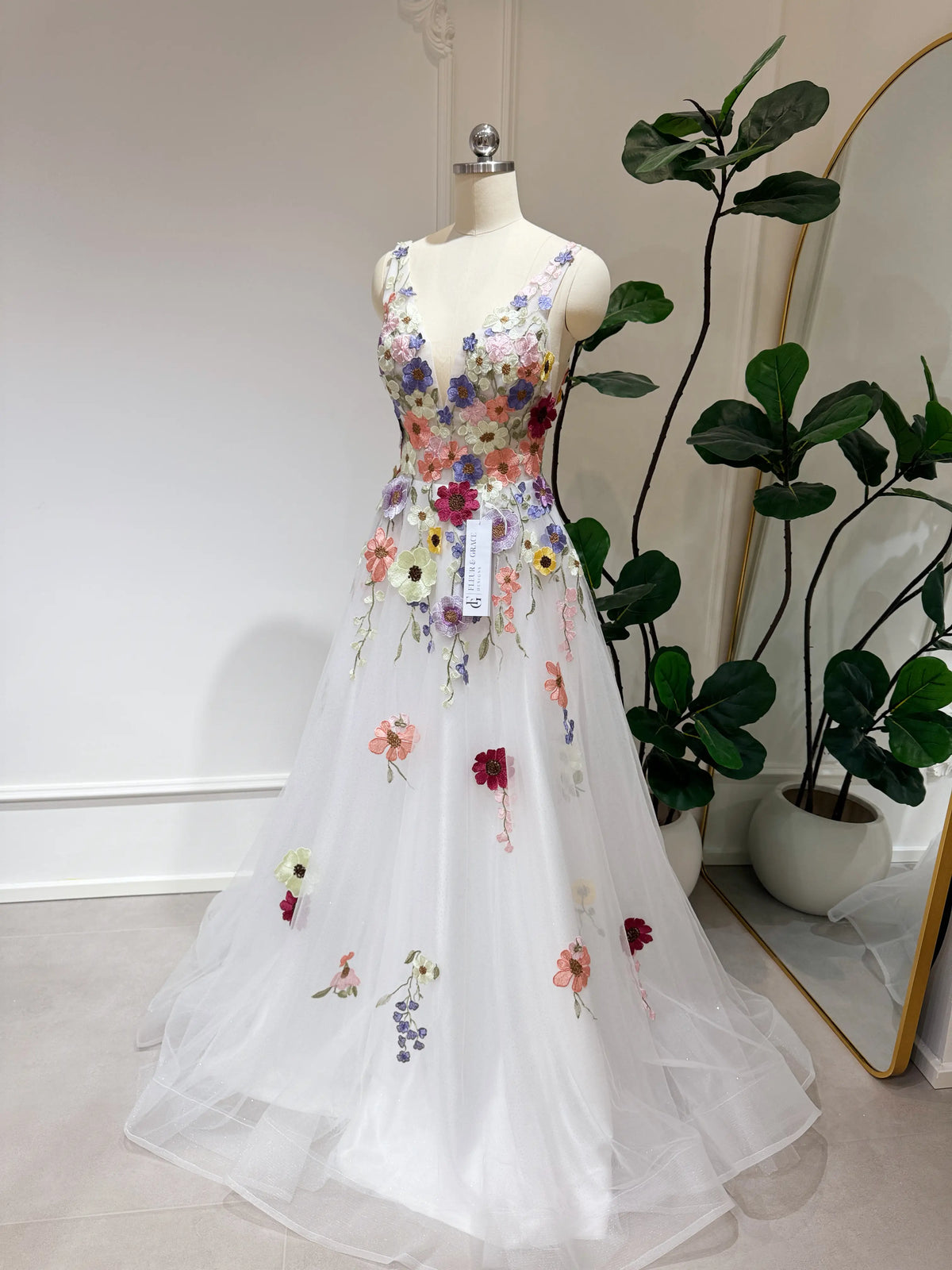 White A-line gown with colorful wildflower embroidery, V-neckline, and sparkly mesh overlay on a mannequin.