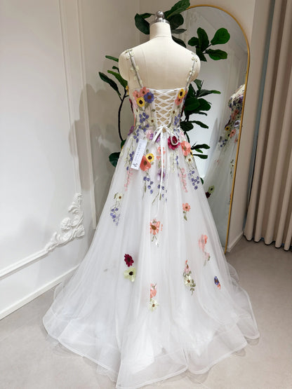White A-line gown with colorful wildflower embroidery, sparkly mesh overlay, and a lace-up back on a dress form.