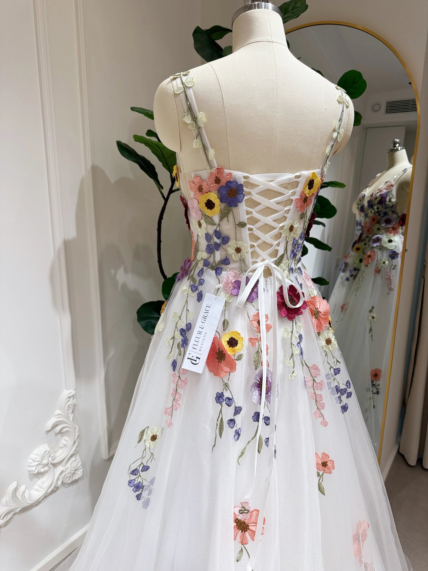 White A-line gown with colorful wildflower embroidery, sparkly mesh overlay, and a lace-up back on a mannequin.