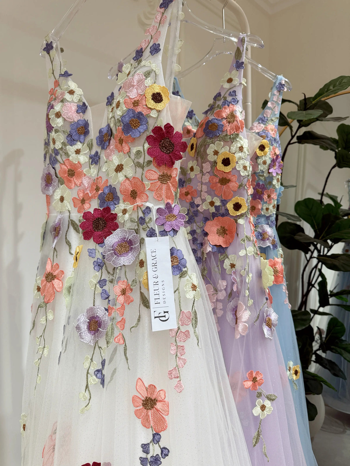 White A-line gown featuring colorful wildflower embroidery on sparkly mesh with a V-neckline and lace-up back.