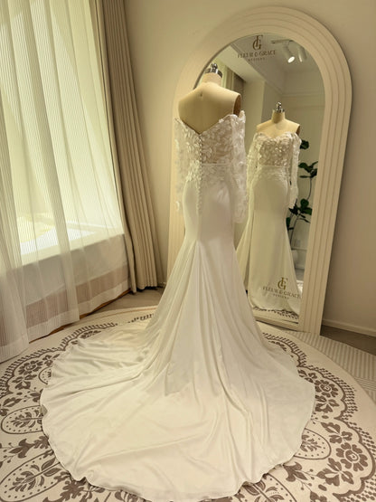 White crepe mermaid wedding dress with off-the-shoulder long sleeves, 3D floral bodice, and a flowing train.