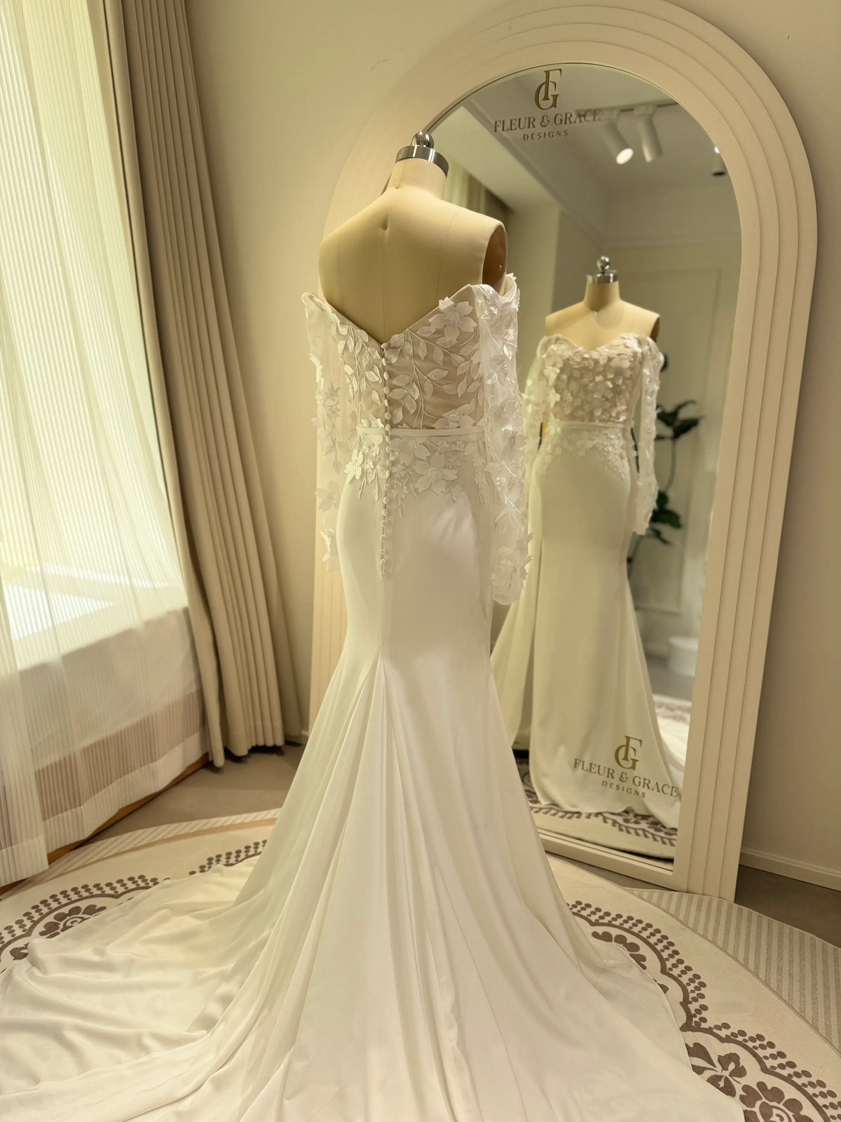 Minimalist white crepe mermaid wedding gown with off-the-shoulder sheer long sleeves and 3D floral bodice details.
